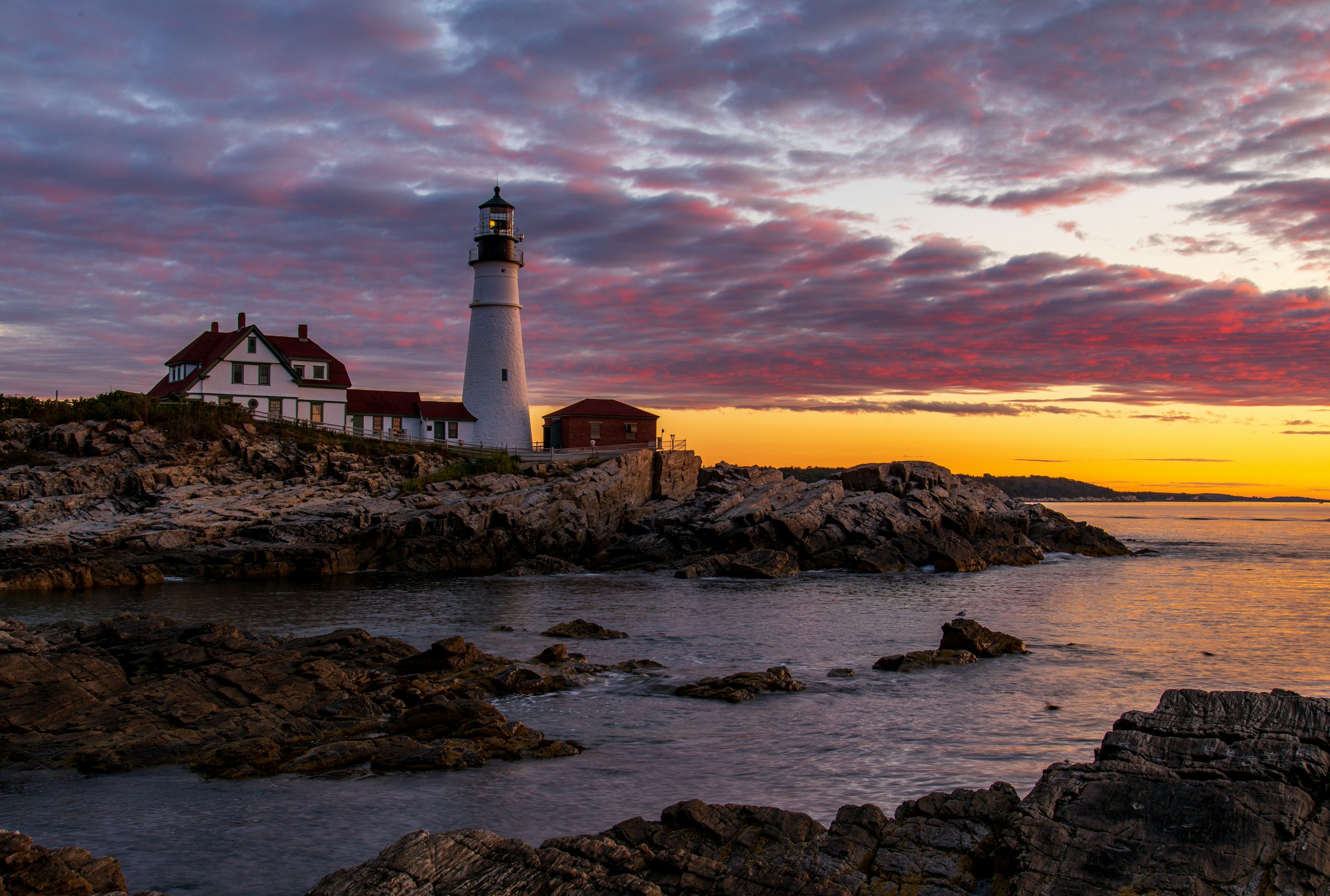 Portland Head Lighthouse at dusk — its lamp lit, the keeper's house beside it, under a violet and amber sky